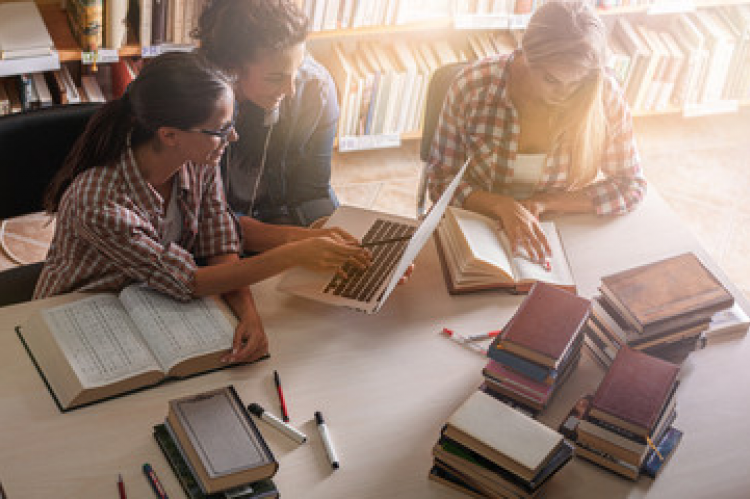 three middle schools students at a table looking at laptop with books around